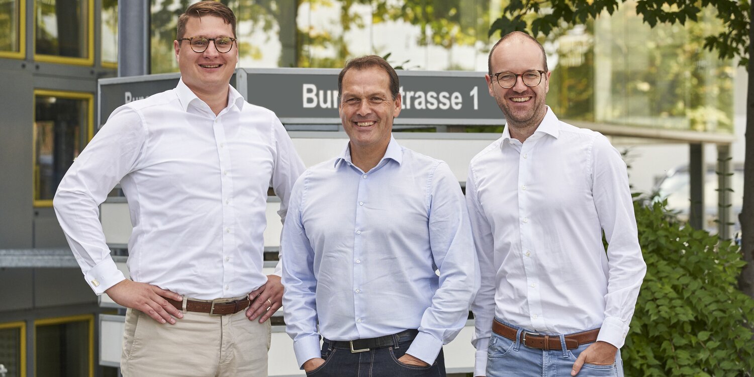 Auf dem Bild stehen drei Männer in weißen Hemden lächelnd nebeneinander im Freien. Hinter ihnen ist ein modernes Gebäude mit gelben Fensterrahmen und Bäumen im Hintergrund zu sehen.