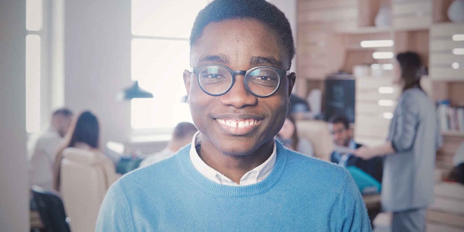 Videostill mit jungem, dunkelhäutigem, lächelnden Mann mit Brille in einem Büro. Im Hintergrund ist eine Arbeitsgruppe sichtbar.