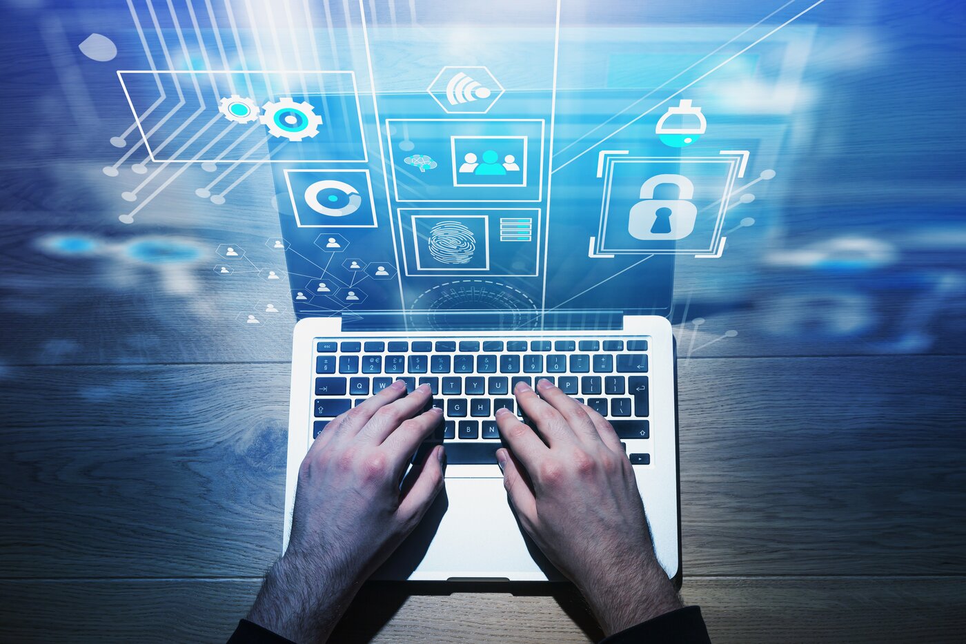 Blick von oben auf Männerhände, die auf einem Laptop auf einem Holztisch etwas tippen. Vor dem Bildschirm fließen Symoble für Cyber-Sicherheit ins Bild. Hintergrund ist verschwommen.