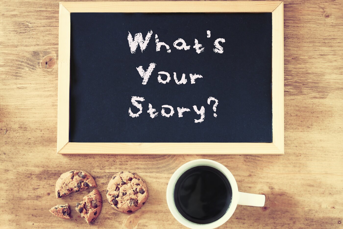 Foto von Schiefertafel auf Holztisch mit geschriebener Frage: What's your Story? Daneben zwei Kekse und eine Tasse Kaffee