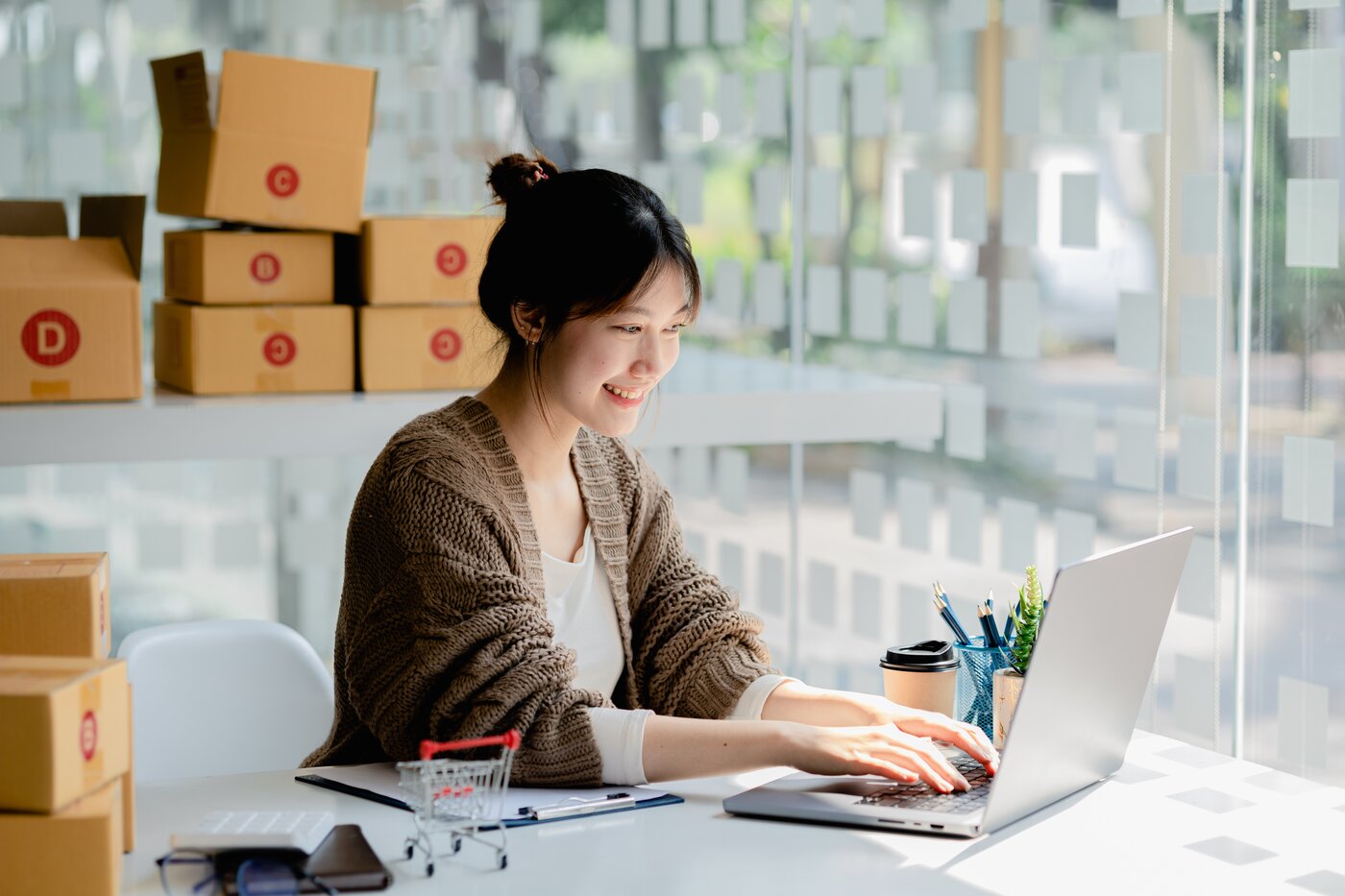 Asiatische Frau am Schreibtisch mit Laptop, im Hintergrund leere Verpackungen und eine Glaswand mit Zetteln